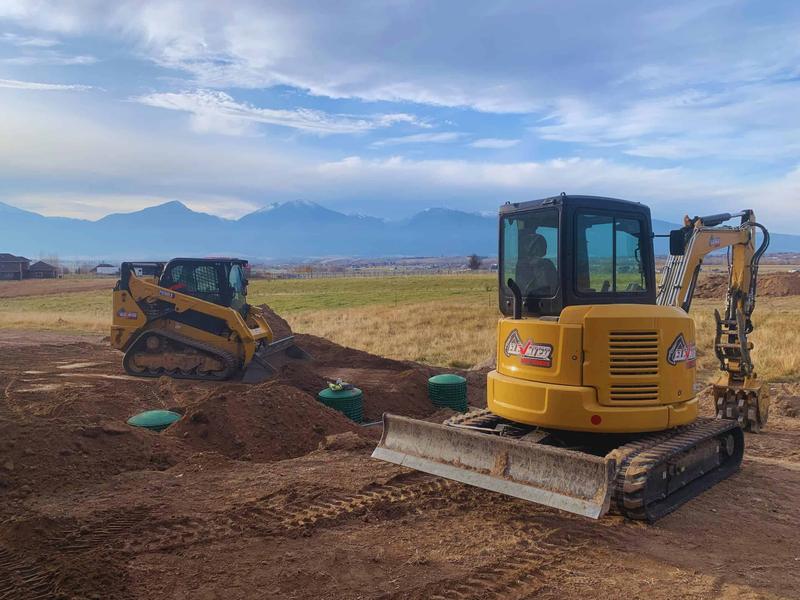 Compact excavator and tracked skid steer on a septic job in the Bitterroot Valley, mountains behind